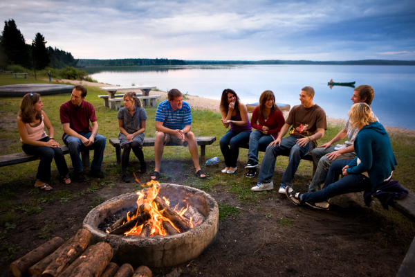Group of friends sitting around a campfire near a lake during dusk in the summertime