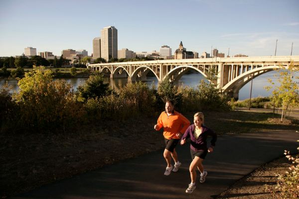 Two people jogging on Meewasin trail with the river and broadway bridge in the background