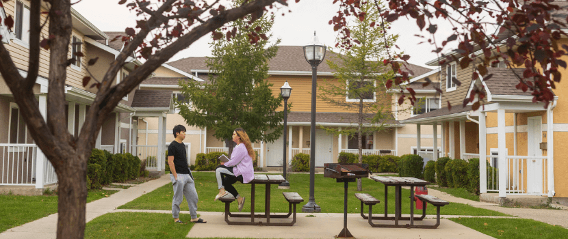Students in the court yard between two sets of housing rows. One student is standing with their hands in their pockets chatting with another student sitting on the top of a picnic table.