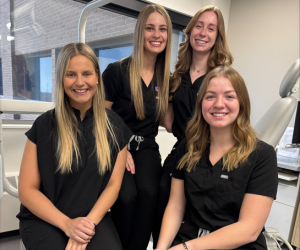 Four students wearing black scrubs sitting on a dental chair smiling at the camera
