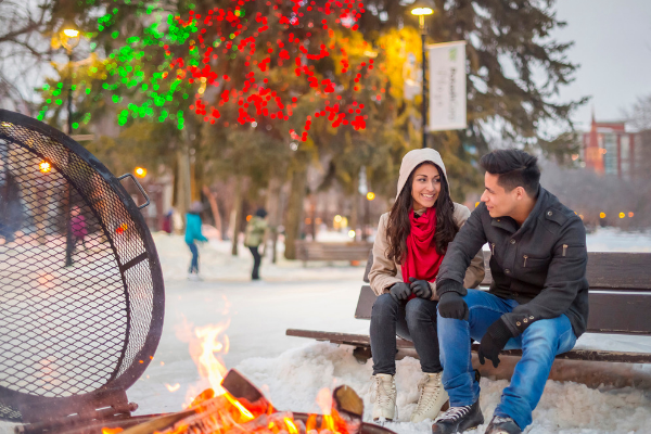 Two people wearing ice skates taking a break on a bench beside a fire at the downtown skating rink