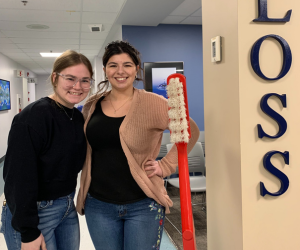 Two students smiling standing next to a large red toothbrush that comes up to one of their shoulders