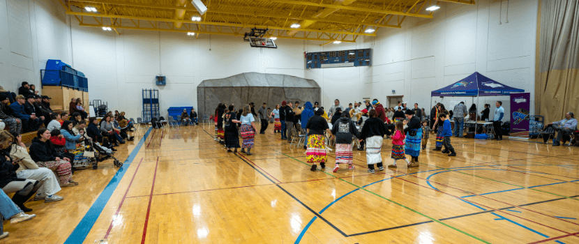 People gathered in the gym with some sitting on bleachers watching dancers and drummers in the middle of the gym