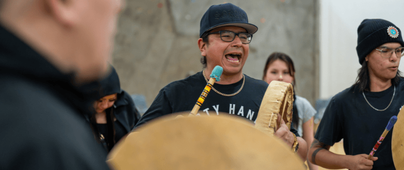 Two people drumming and singing with people dancing in the background